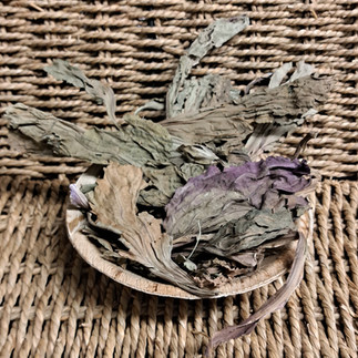 A selection of large greeny/purple broadleaf plantain leaves in a round palm leaf bowl in front of a light brown wicker background