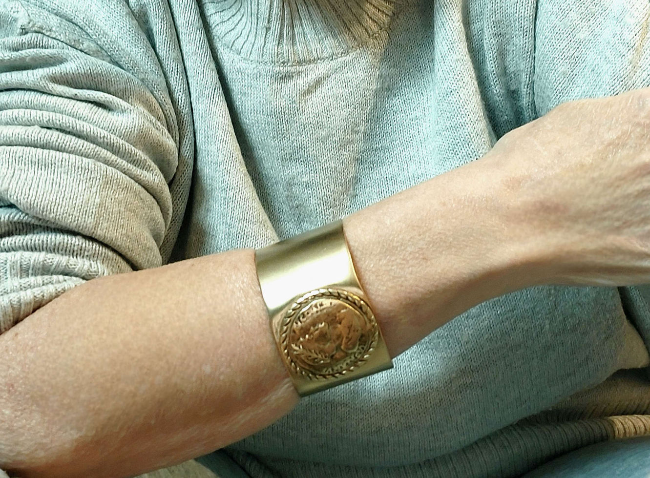 יד עם צמיד זהב גדול, יפהפה, על רקע סוודר אפור ולבן.