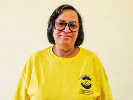 Carolyn is standing against a plain light-colored wall, wearing dark-framed glasses and a bright yellow T-shirt. The shirt features a small logo on the left chest with a handshake graphic and the text “New Avenues to Independence, Inc.” The person is centered in the image, facing the camera, with short dark hair and a simple indoor background.