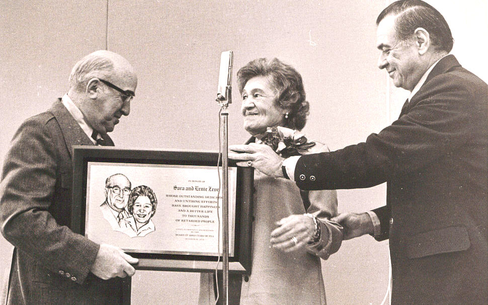 Man presents framed award plaque to a woman, microphone nearby.