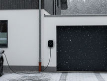Ein modernes Einfamilienhaus im Winter mit einer angeschlossenen Wallbox an der Hauswand. Ein weißes Elektroauto ist davor geparkt und wird geladen. Schnee bedeckt den Boden und das Auto – Symbolbild für Elektromobilität und nachhaltige Energieversorgung auch bei winterlichen Bedingungen.