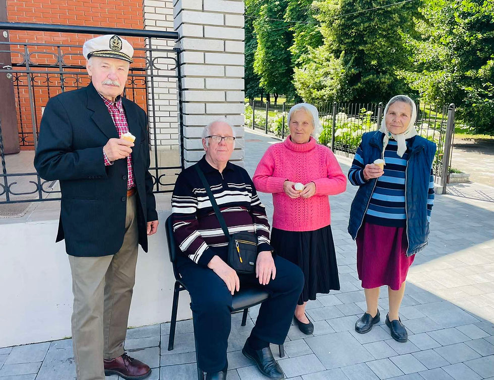 Two women stand on a paved walkway holding food. One wears a pink sweater; the other wears a blue-striped shirt. Greenery in the background.
