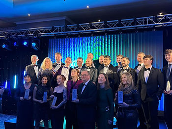 A group of people in formal wear stand on a stage holding awards. The background features blue lights, and the mood is celebratory.