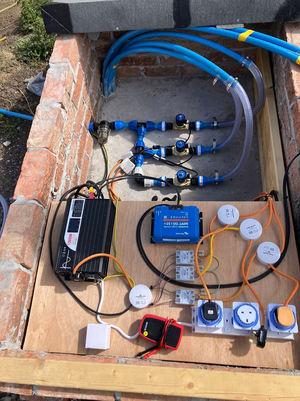Open utility box with blue hoses, valves, inverter, wiring, and outlets on a plywood base beside brick walls, outdoors