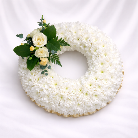 Round funeral wreath of white chrysanthemums with cream roses and greenery accent, displayed on a soft white and grey silk background