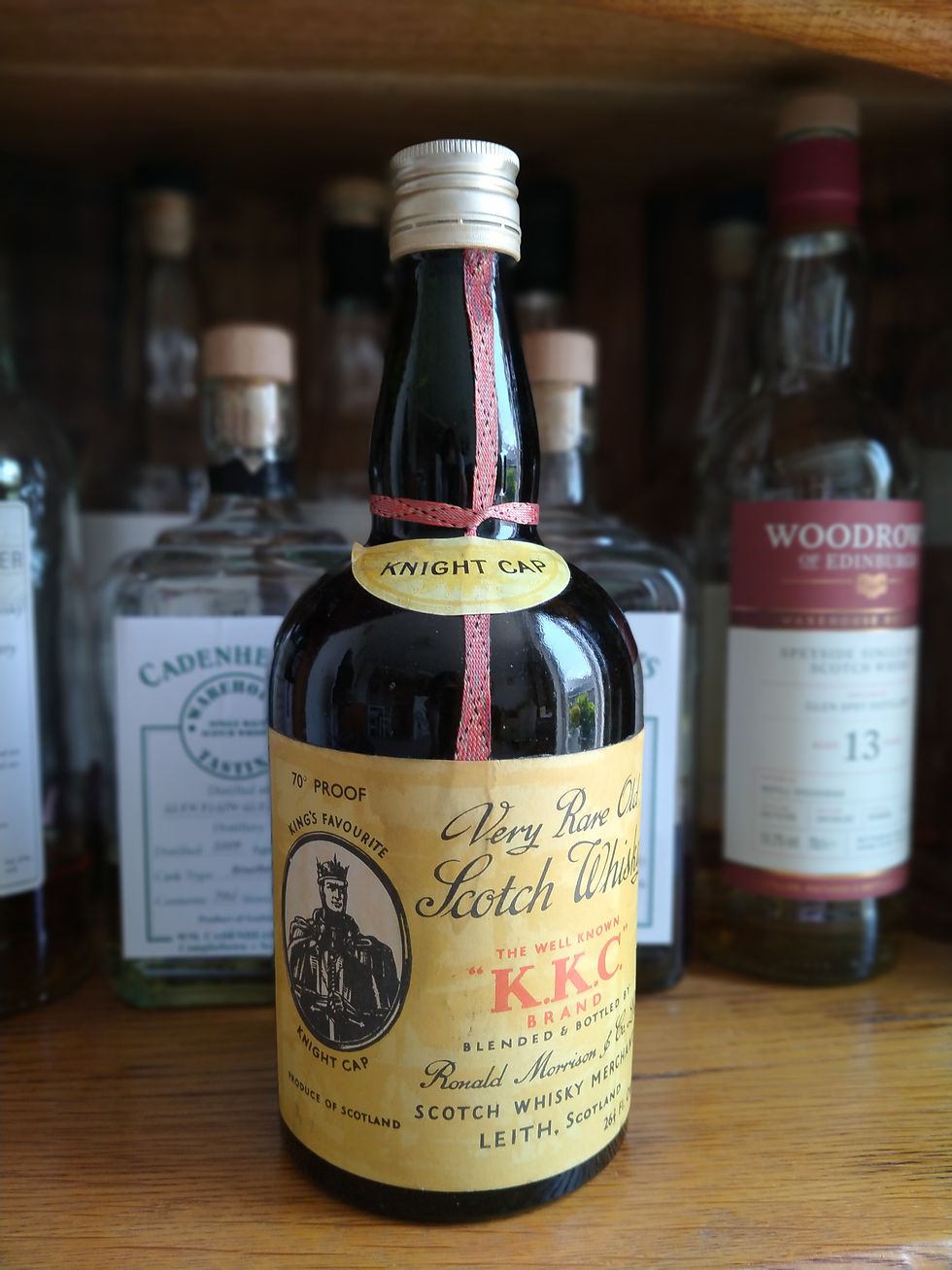 Vintage Scotch whisky bottle labeled Knight Cap and K.K.C. on a wooden shelf, with other whisky bottles blurred behind it.