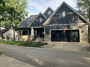 A dark gray craftsman-style house with stone accents, a two-car garage, and an American flag hanging by the front entrance.