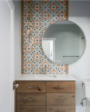 A modern bathroom featuring a wooden vanity with a white sink, topped by a patterned tile panel and a large round mirror. Delicate pendant lights hang in front of the tiles.