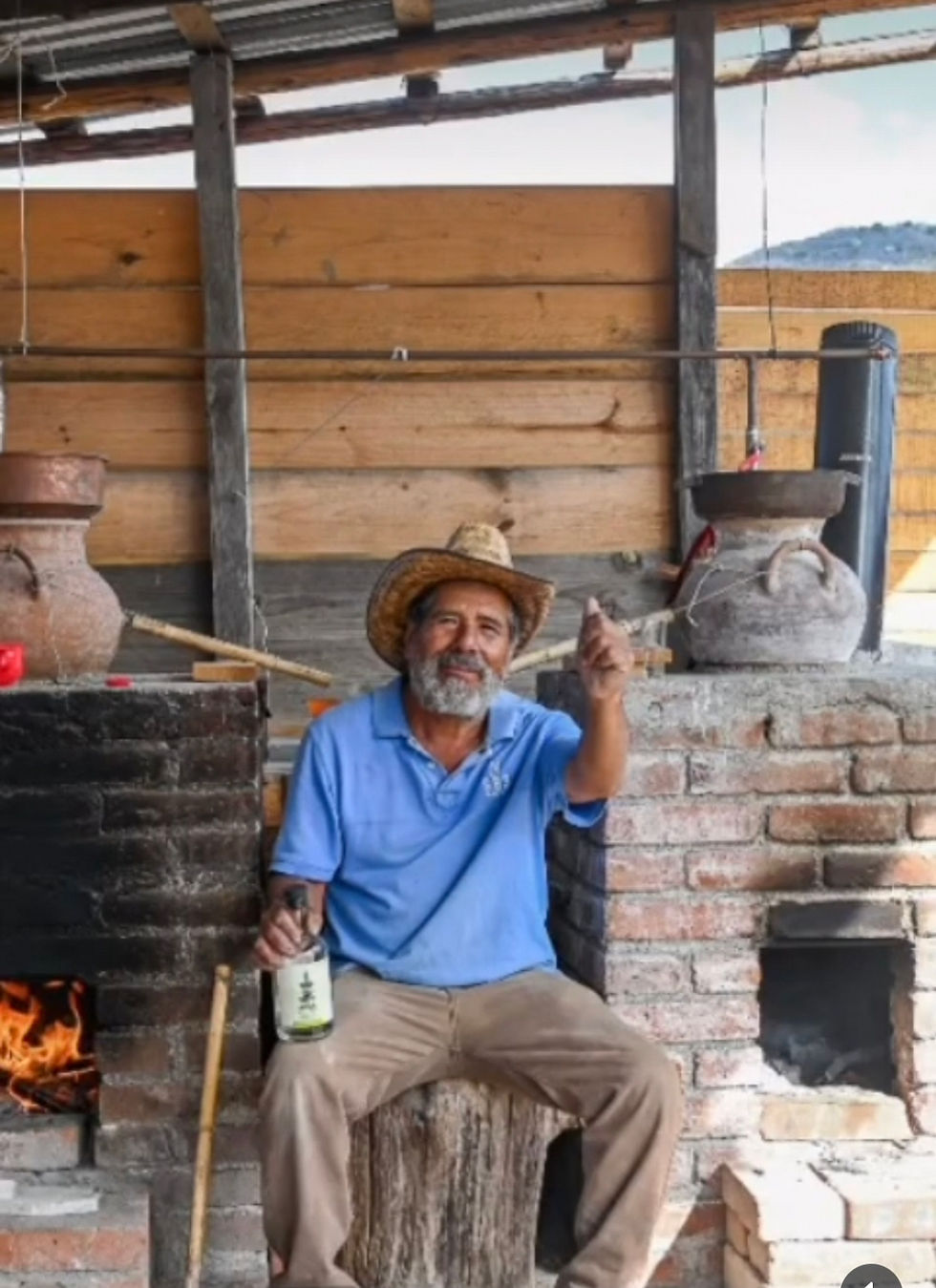 Don Ignacio Gutierrez sitting beside a clay still and fire pit, holding a bottle of Con Todo El Alma Mezcal Ancestral at his traditional palenque in Santiago Matatlán.