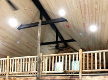 A loft with wood railings beneath a cathedral ceiling with dark beams and a ceiling fan.