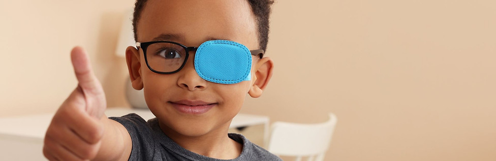 Ein Junge mit Augenklappe auf der Brille zur Behandlung von Strabismus zeigt im Zimmer den