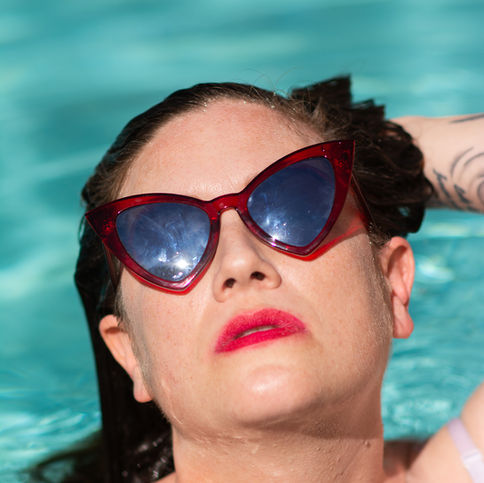 Close up of Dahlia Snow a brunette femme presenting dominatrix, in a pool with red lip stick and red cat eye sun glasses on