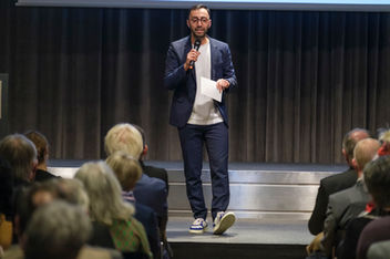 Mann spricht mit Mikrofon auf Bühne zu Publikum bei einer Präsentation