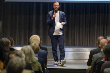 Mann spricht mit Mikrofon auf Bühne zu Publikum bei einer Präsentation