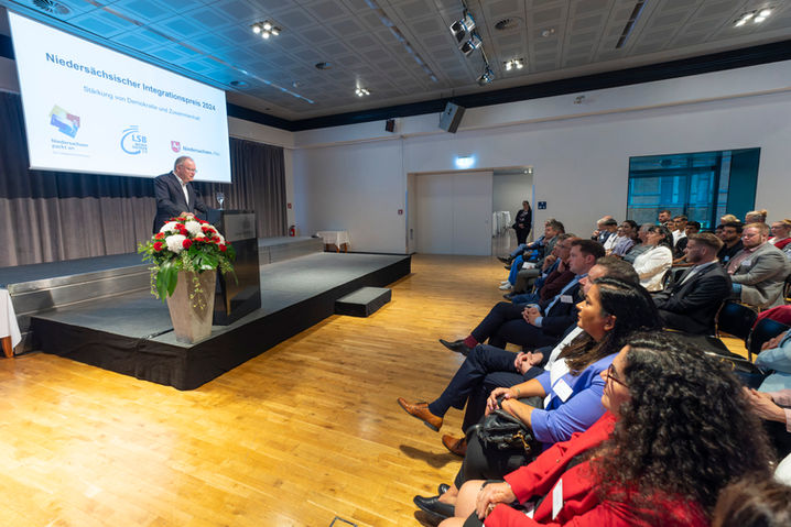 Redner steht auf der Bühne und spricht vor Publikum. Text auf der Leinwand.