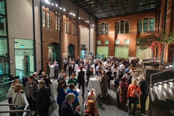 Crowd gathers at an event, people standing and chatting indoors with bright lights.