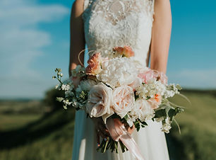 A bridal bouquet made of pink roses, white mum and other filler flowers. Bride is holding bouquet at her waist while standing in a field.