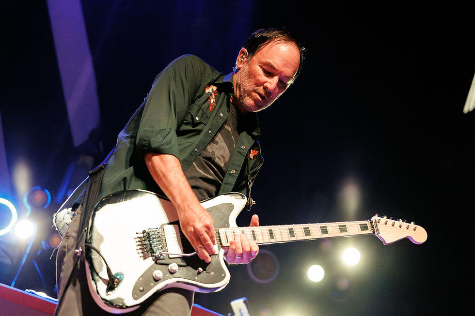Musician playing a white electric guitar on stage, wearing a dark shirt with floral details. Bright spotlights in the background.