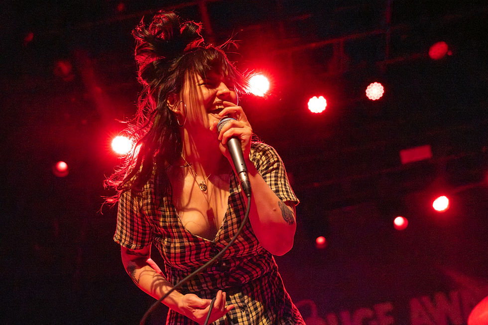 A woman passionately sings into a microphone on stage, under dramatic red lighting. She wears a plaid dress, with text partially visible behind her.