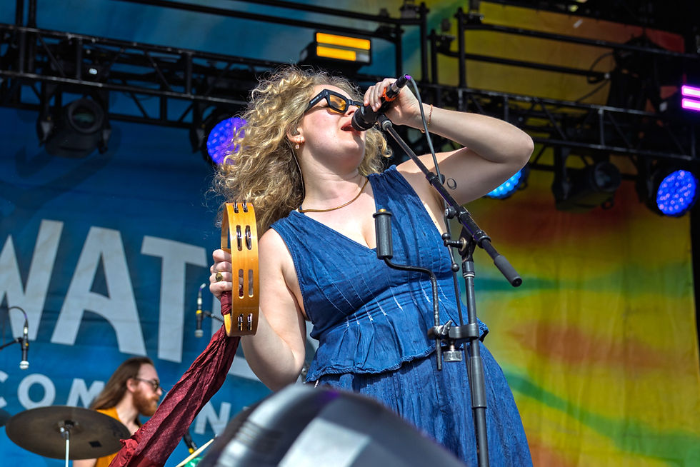 A woman with curly hair sings into a microphone, holding a tambourine on stage. She wears a blue dress, with colorful lights in the background.