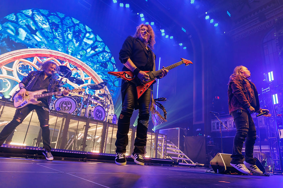 Rock band performing energetically on stage with colorful lights. Guitarist in black with red guitar, blue patterned backdrop, intense atmosphere.