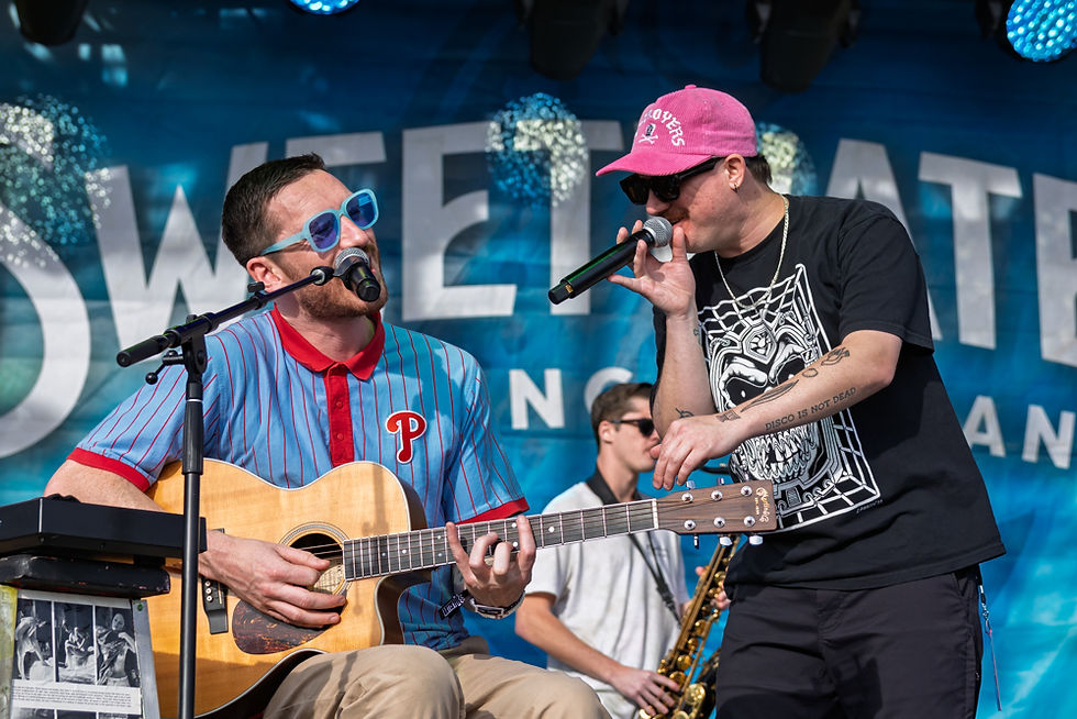Two musicians perform on stage. One plays acoustic guitar and wears blue sunglasses, the other sings into a mic. Blue "Sweetwater" banner behind.