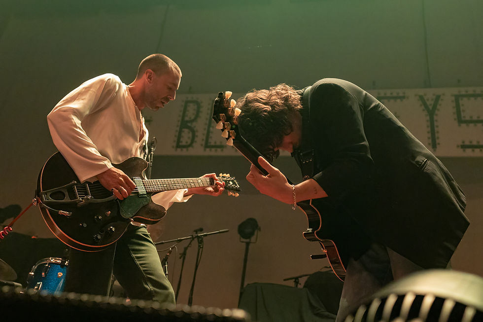 Two guitarists performing on stage, leaning forward passionately. Dim lighting, drums in the background. Sign reads "BL" partially visible.