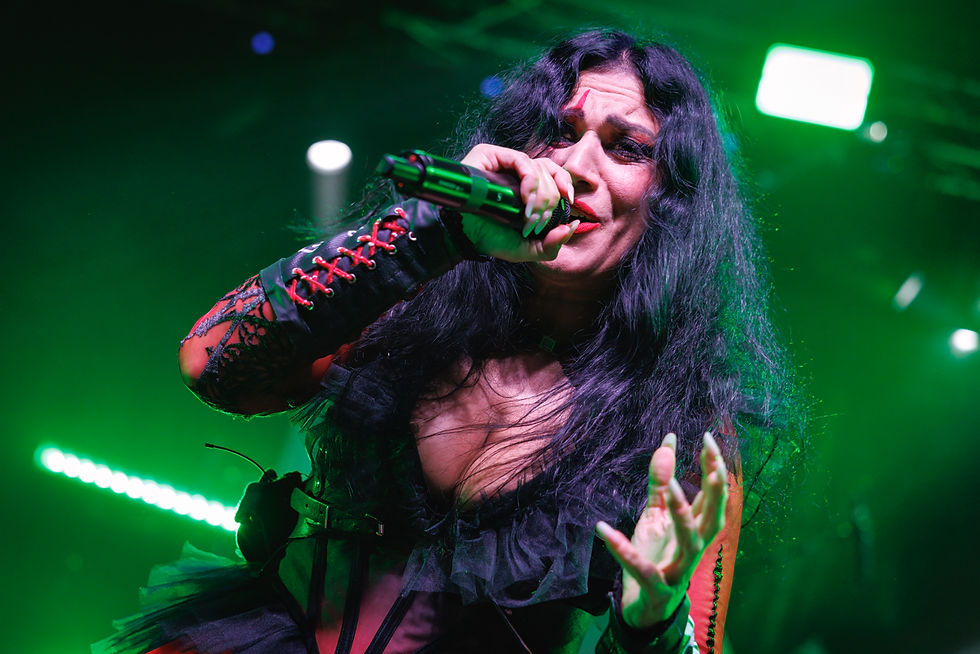 Singer with long black hair, wearing a black and red outfit, passionately performs on stage under green lights, holding a microphone.