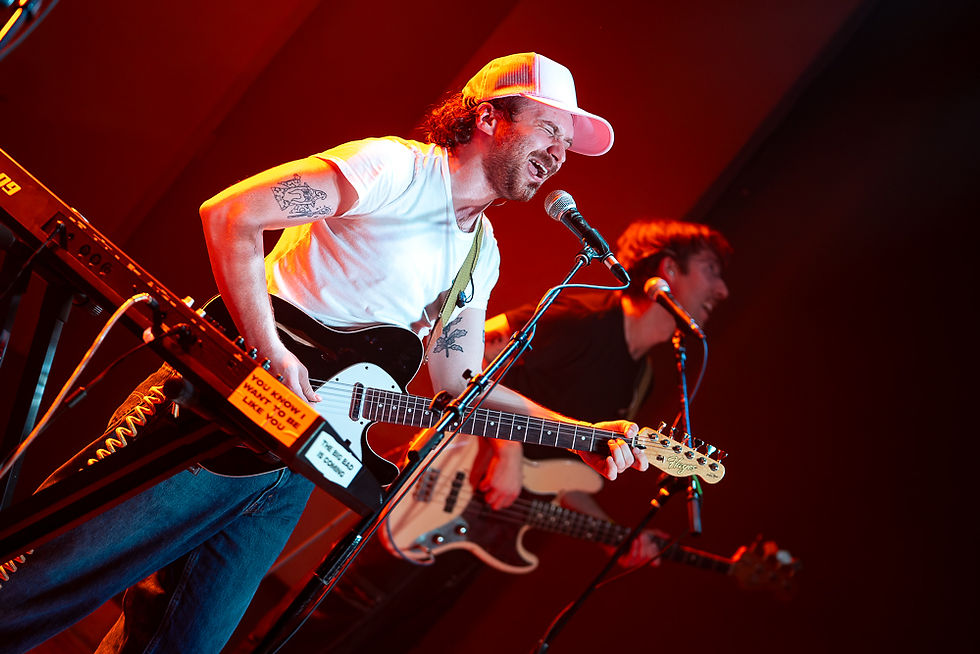 Two musicians perform energetically on stage. One sings into a mic, playing a black and white guitar, wearing a white shirt and cap. Red lighting.