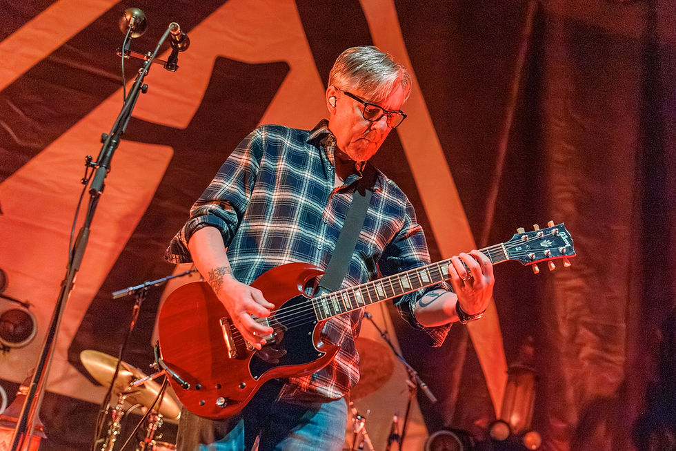 Musician in plaid shirt plays a red electric guitar on stage. Microphone nearby. Background features abstract shapes. Energetic mood.