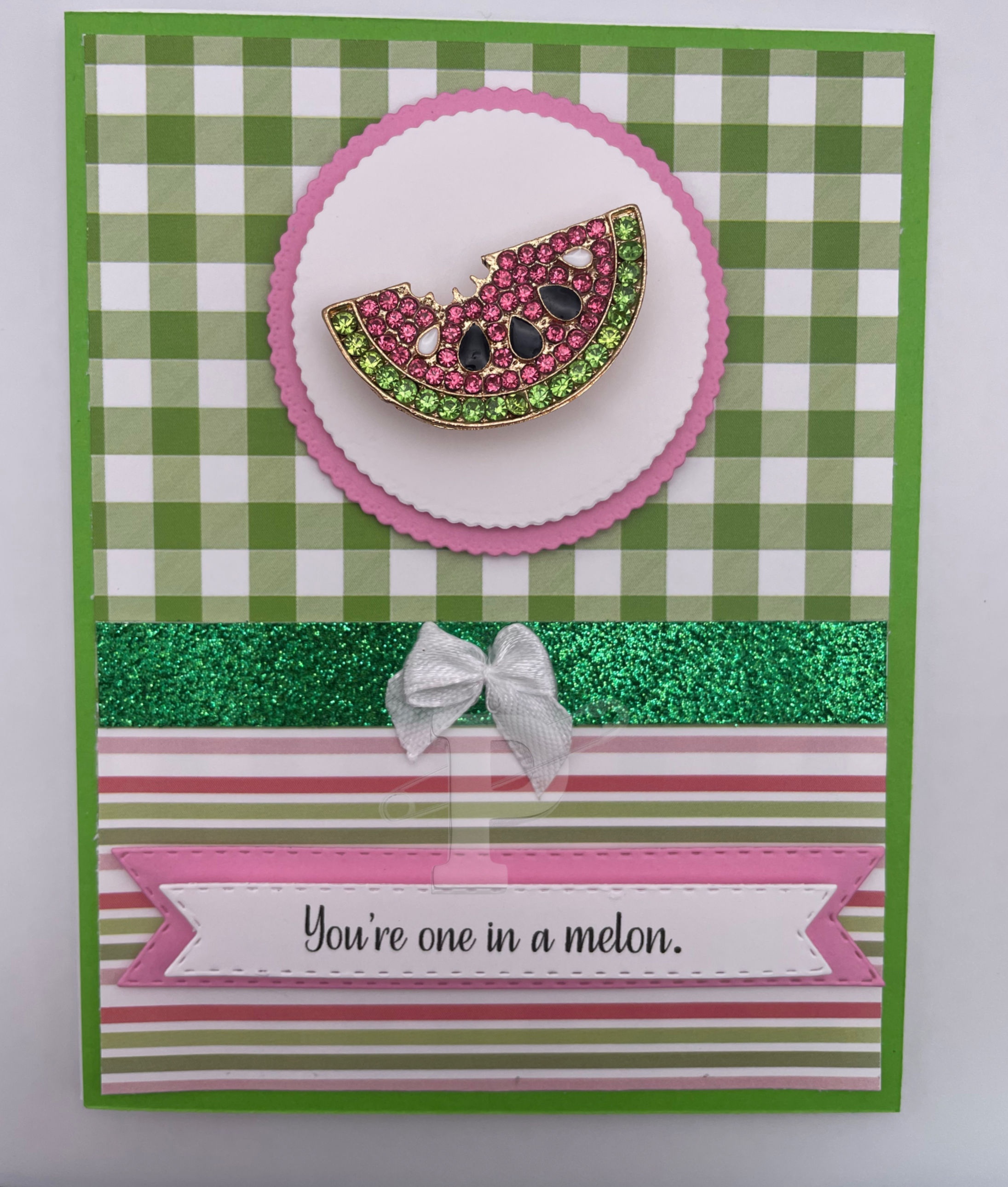 Greeting card with rhinestone watermelon slice brooch on green gingham background; features "You’re one in a melon."