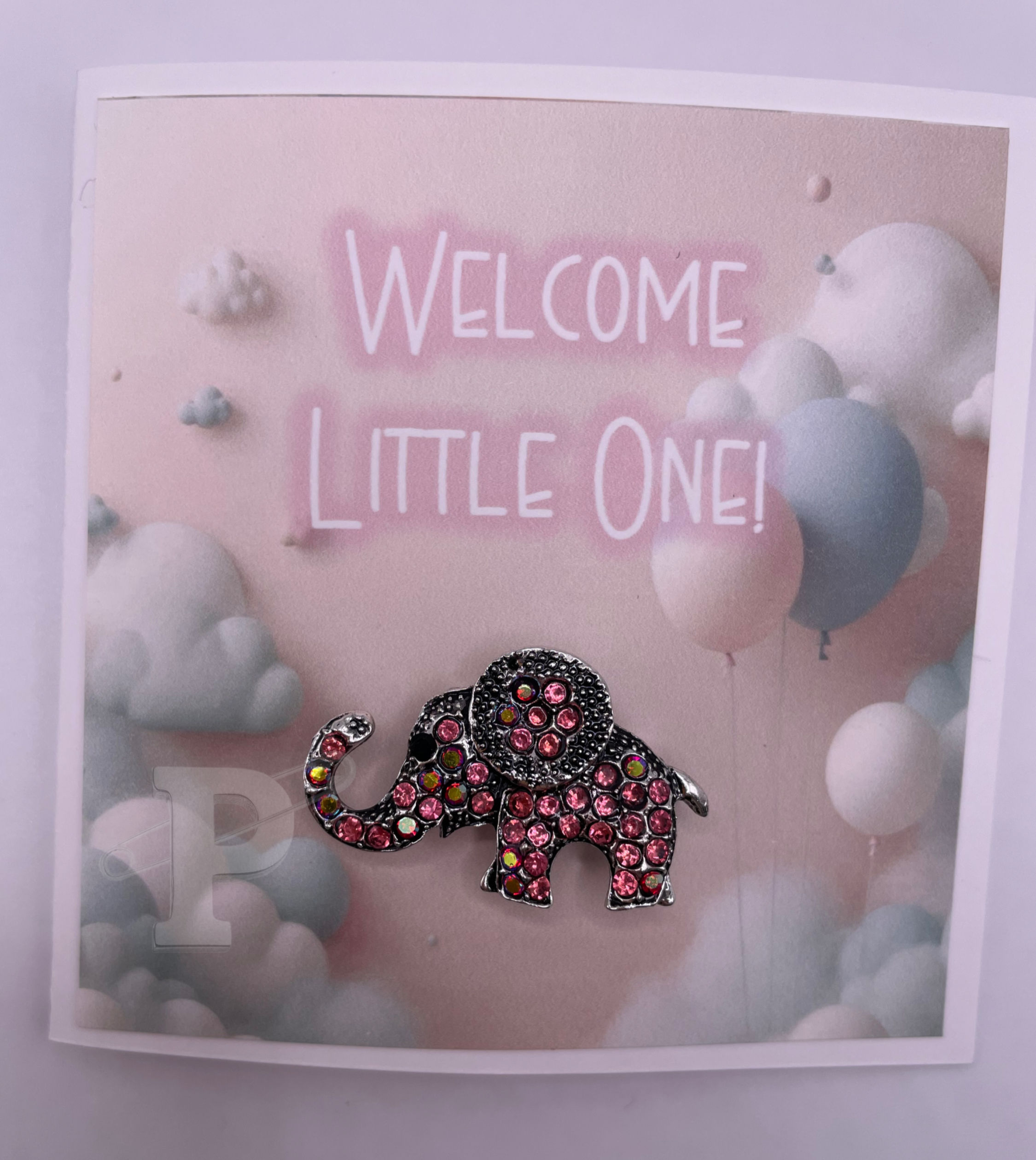 Mini card that reads “Welcome Little One!” with pink rhinestone elephant brooch on soft pastel cloud and balloon background.