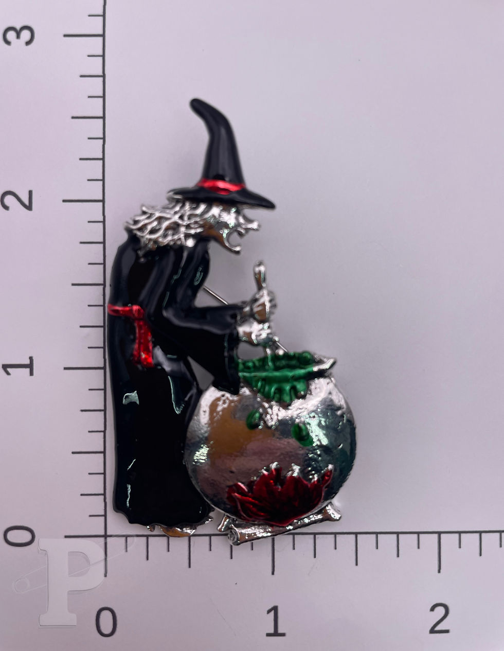 Thumbnail: Enamel witch and bubbling cauldron brooch with silver, black, red, and green accents shown with ruler for size reference.