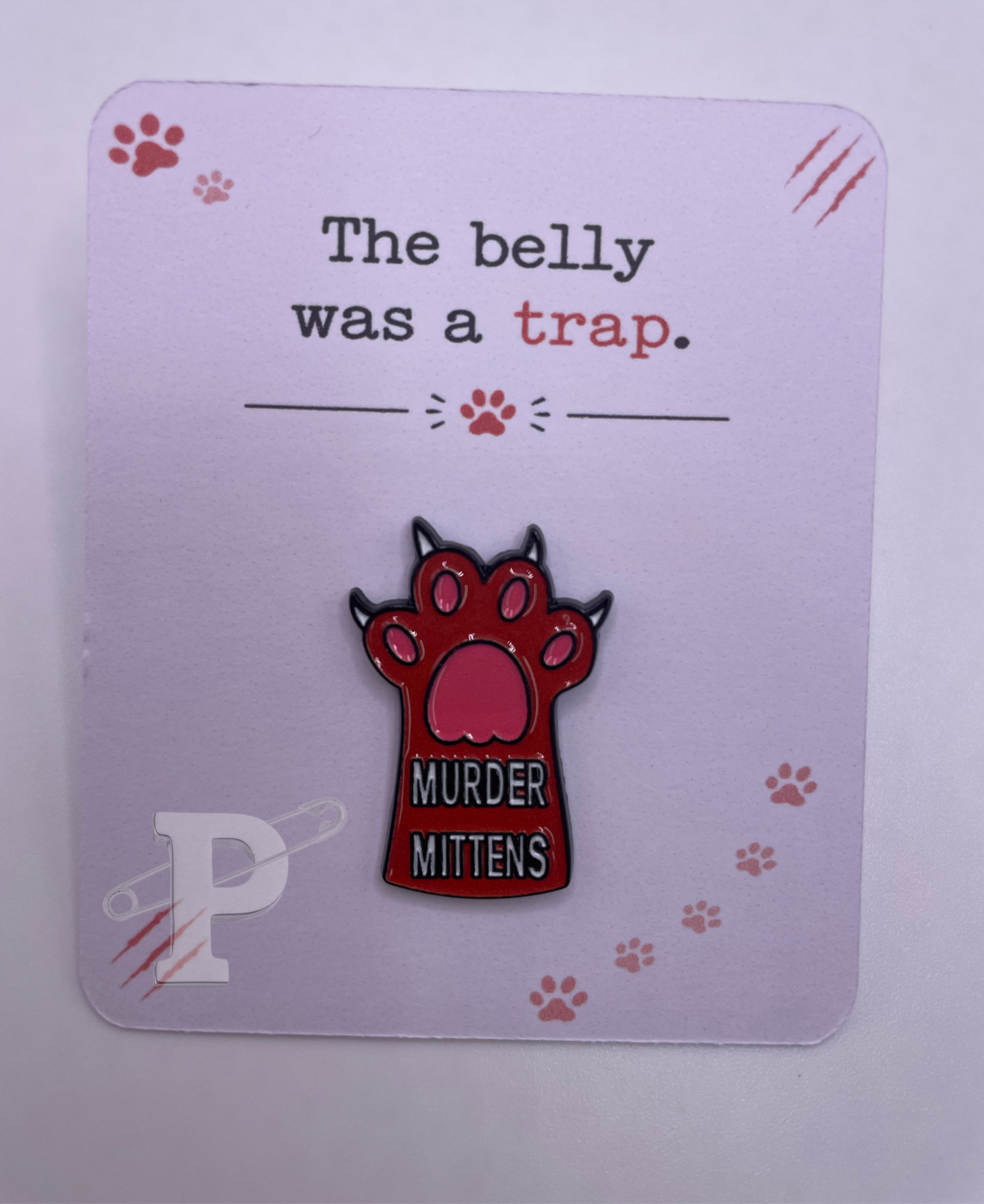 Murder Mittens cat paw enamel pin displayed on a playful paw-print card with the sentiment “The belly was a trap.”