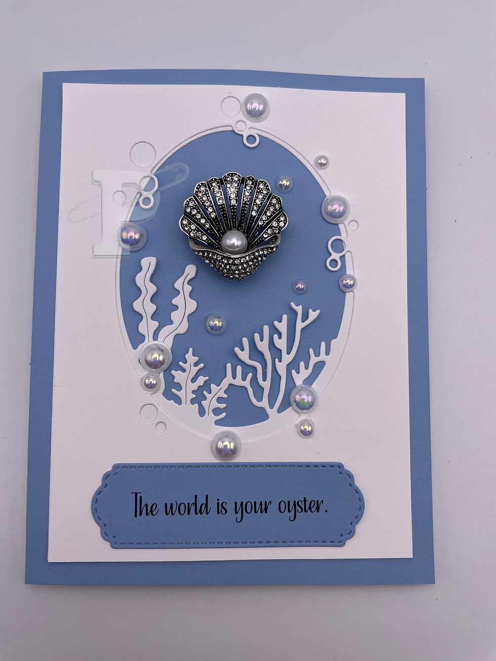 Elegant greeting card with silver oyster brooch, set against a deep sea with the sentiment "The world is your oyster".