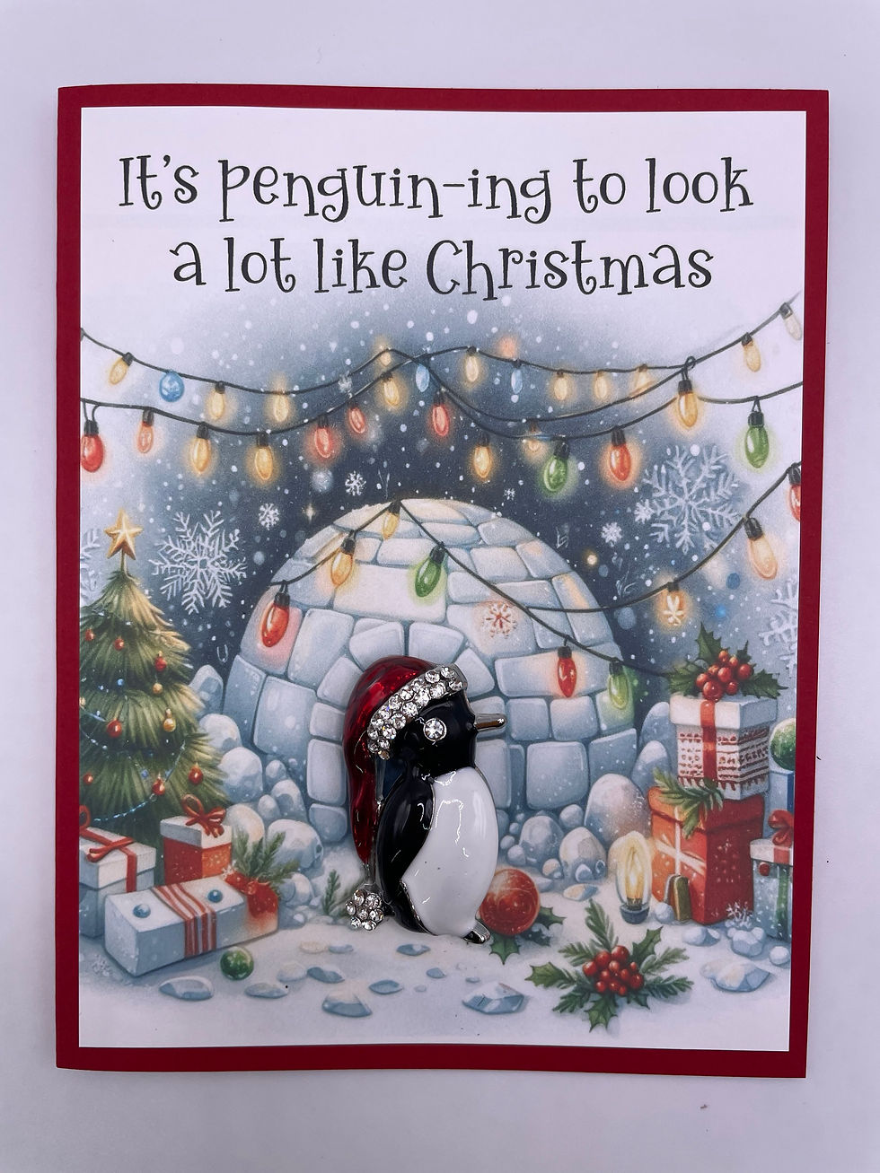 Christmas card with penguin brooch wearing a Santa hat, set against an igloo decorated with lights, presents, and a tree