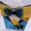 Thumbnail: Top view of suit jacket-shaped card with peacock blue background, gold and teal feather bow tie, and matching feather pin