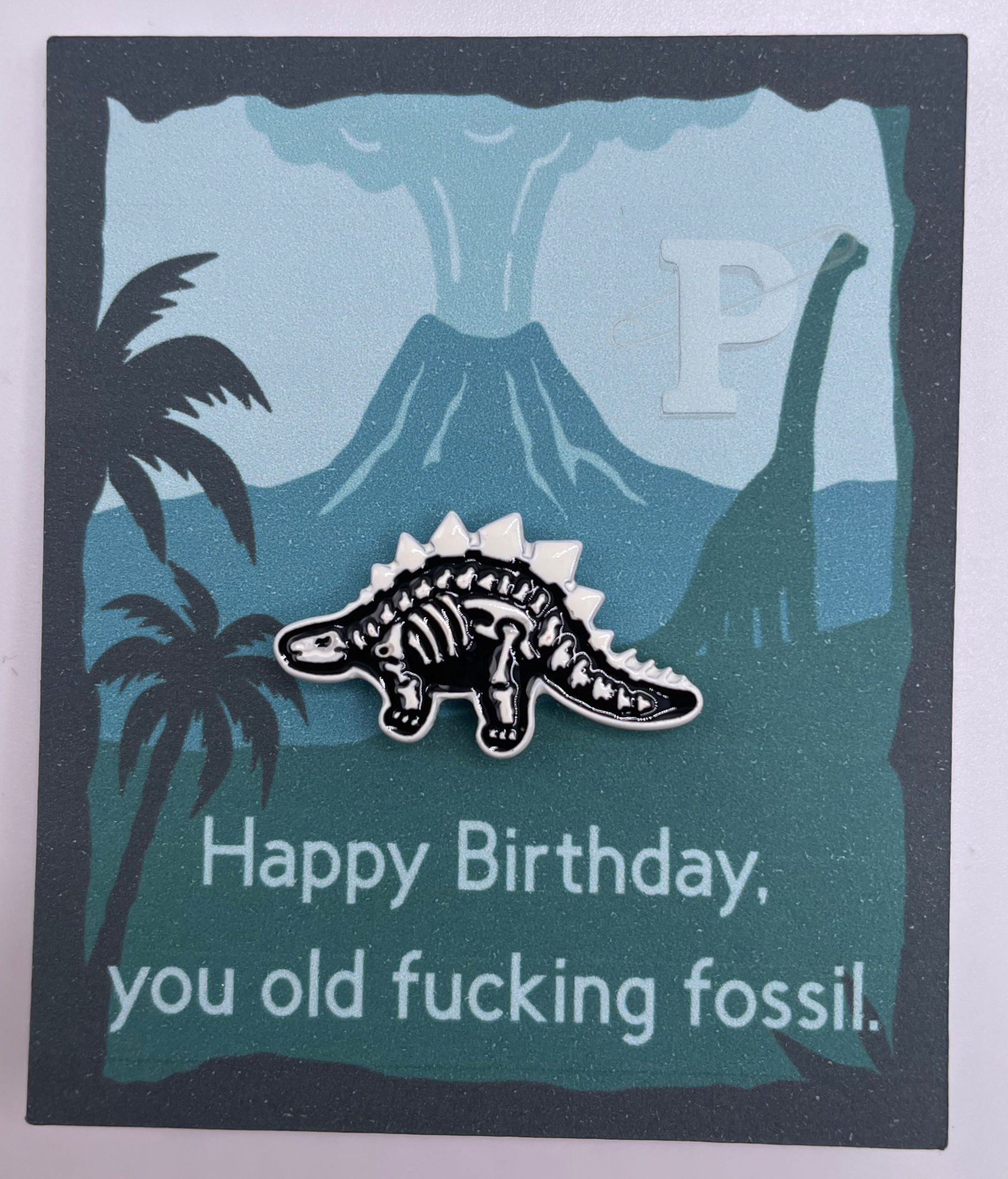 Flat card that reads “Happy Birthday, you old fucking fossil.” with dinosaur skeleton lapel pin.