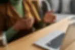Person gesturing beside a laptop on a wooden desk in a cozy office, wearing green and brown, mid-conversation.