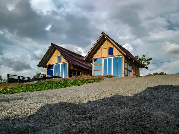 Two modern wooden A-frame cabins with large glass windows sit on a sandy hillside under a cloudy sky. The cabins have metal roofs and a simple, rustic design, blending with the natural surroundings.webp