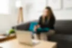 Woman on a gray couch talks on a laptop in a bright living room, with teal pillows and wall art reading Learn and Live.