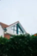 A house with a brick facade, large glass windows, and a triangular roof is partially obscured by lush green shrubs under a cloudy sky.