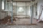 Room under renovation with debris on the floor, metal supports, an open window, ladder, and tools against white walls. Bright atmosphere.
