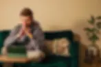 Pensive man in a gray hoodie sits on a green sofa, hands clasped, looking at a tablet; plant and cozy decor nearby.