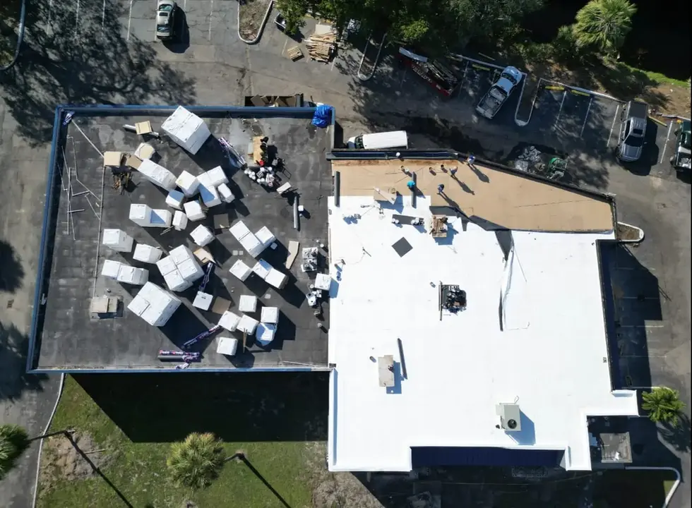Overhead drone image showing two connected commercial roofs, one completed in white membrane and one under construction with materials and workers present