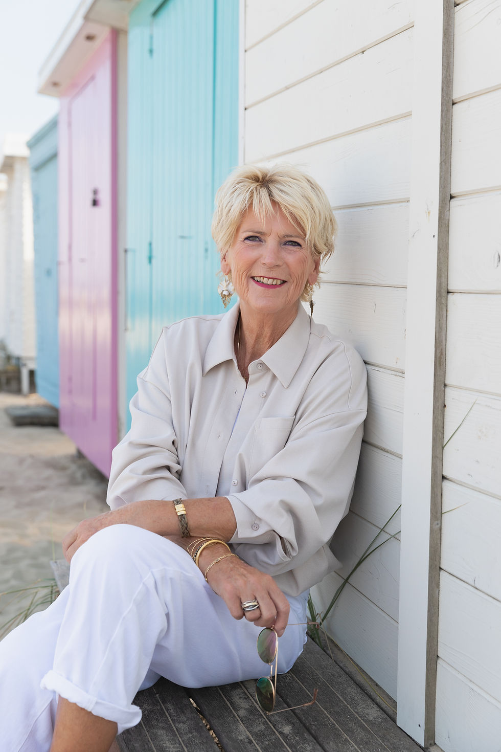 Lachende blonde vrouw zit tegen witte houten wand bij pastelkleurige strandhuisjes en houdt een zonnebril vast.