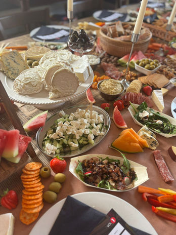 Grazing table with assorted cheeses, fruits, crackers, dips, cured meats, olives, vegetables, nuts, and lit candles — arranged for a rustic party setting.