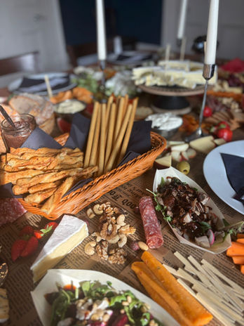 Grazing table with breads, cheeses, cured meats, fruits, dips, crackers, vegetables, olives, nuts, and lit candles arranged for a rustic party spread.