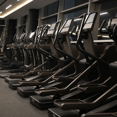 Row of treadmills in a modern gym with dim lighting. Large windows line the wall, and stairs lead to an upper level. Mood is calm.