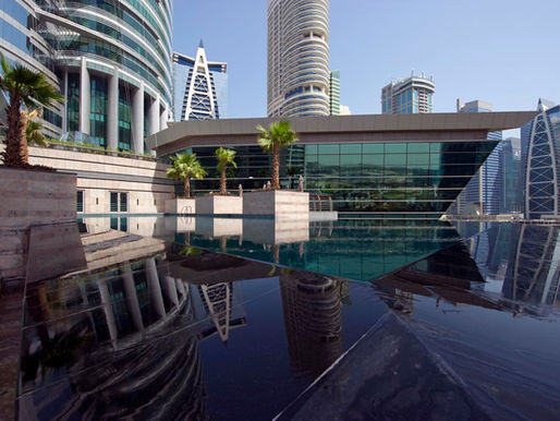 Modern skyscrapers with mirrored glass reflect in calm water, surrounded by palm trees. Urban cityscape under a clear blue sky.